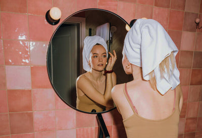 Image montrant une femme avec une serviette sur la tête, appliquant sa routine beauté devant un miroir rond dans une salle de bain aux carreaux roses