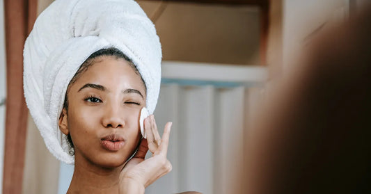 Femme avec une serviette sur la tête appliquant un soin avec un coton sur son visage, routine de nettoyage pour peau grasse.
