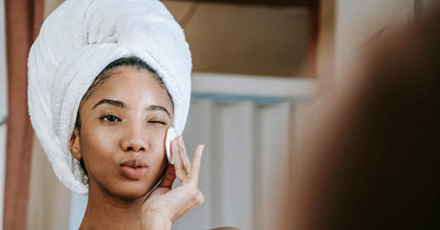 Femme avec une serviette sur la tête appliquant un soin avec un coton sur son visage, routine de nettoyage pour peau grasse.